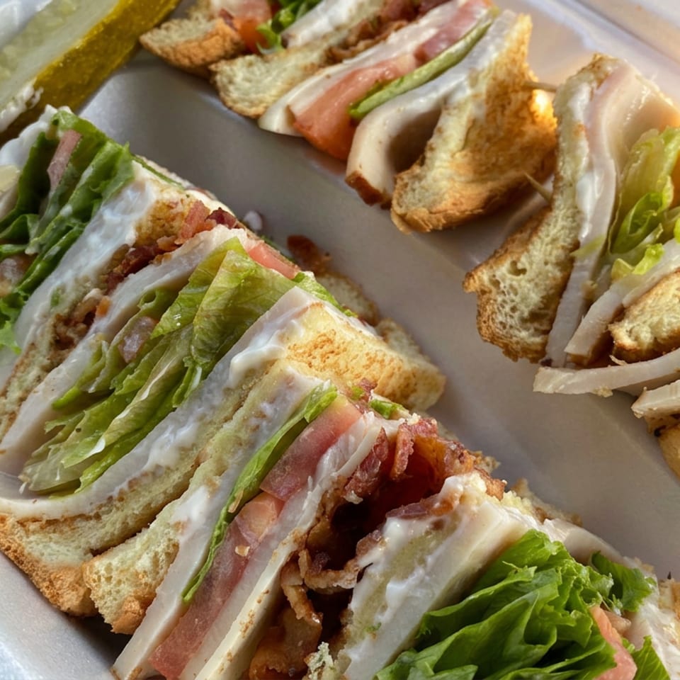 Assorted club sandwiches with turkey, bacon, lettuce, tomato, mayonnaise, and pickles arranged in a foam takeout tray.